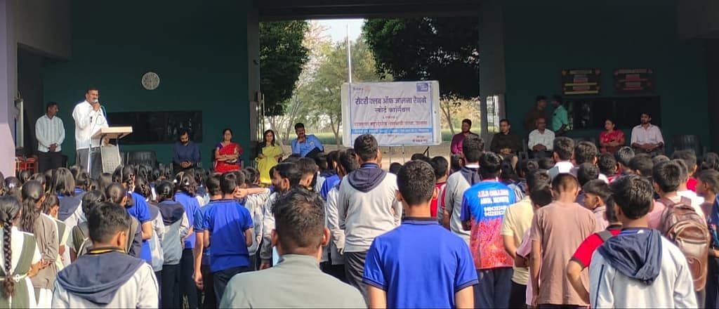 WhatsApp Image 2026-02-15 at 163327 - NewsNation Online A large group of students gathered in an open space, listening to a speaker at a podium. The setting features a banner and several adults sitting at a table in the background, with trees visible outside.