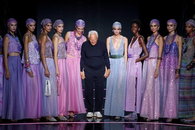 images-21 - NewsNation Online A group of models wearing elegant purple and pink gowns stands alongside a designer holding hands with one of them, all appearing on a fashion runway.